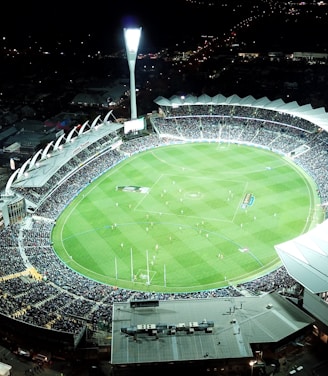 An aerial view of a large, illuminated sports stadium filled with spectators. The stadium is oval-shaped, featuring a green field in the center with markings for a sports game, likely football or rugby. Bright floodlights illuminate the scene, creating a vibrant atmosphere. Surrounding the stadium are urban buildings and streets, barely visible in the night.
