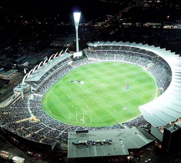 An aerial view of a large, illuminated sports stadium filled with spectators. The stadium is oval-shaped, featuring a green field in the center with markings for a sports game, likely football or rugby. Bright floodlights illuminate the scene, creating a vibrant atmosphere. Surrounding the stadium are urban buildings and streets, barely visible in the night.