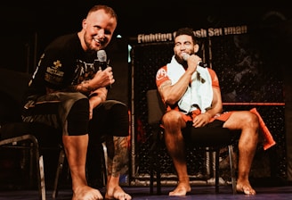 Two individuals are seated on a stage, each holding a microphone and smiling. They are in a sports-related setting with one wearing a casual black outfit and the other in sportswear, with a towel around his neck. The background includes elements of a fighting or martial arts cage.