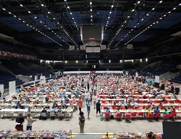 An indoor sports arena filled with rows of tables, each occupied by people engaged in competitive activities. The ceiling is high, with numerous lights illuminating the large space. The audience seating on either side is empty, and there are flags hanging from the upper level.