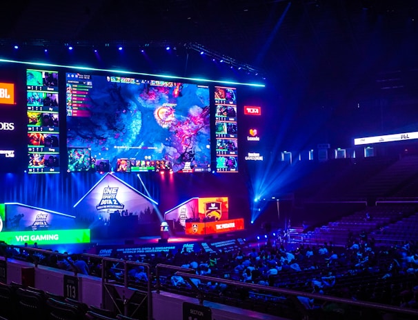 A large esports arena featuring a massive screen displaying a video game, surrounded by sponsor logos and team names. The auditorium is filled with spectators watching the gaming event. Neon blue and purple lights illuminate the stage, creating a vibrant atmosphere.