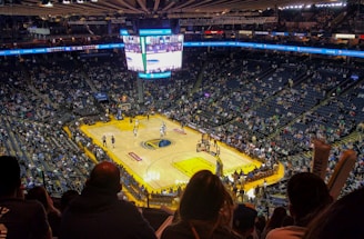 An indoor basketball arena filled with spectators. The court features a central logo and is surrounded by partially occupied tiered seating. A large scoreboard hangs above the center of the court, displaying the game score and time. Ambient lighting highlights the players on the court and the vibrant atmosphere of a live sports event.