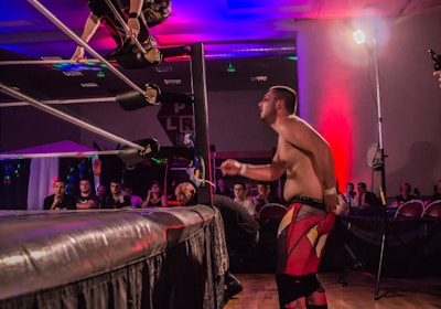 A wrestling match taking place in an indoor venue, where one participant is crouched on the top rope of the ring, while the other stands outside the ring looking at them. A crowd watches the event enthusiastically from their seats.
