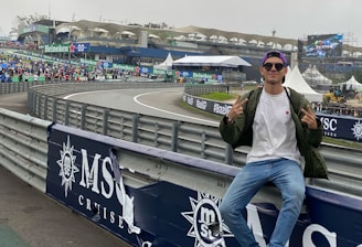 A person in casual attire sits on a barrier beside a racetrack, making peace signs with both hands. In the background, there is a stadium, a large crowd, promotional banners, and a screen displaying an advertisement. The scene has an overcast sky, and the setting appears to be a motorsport event.