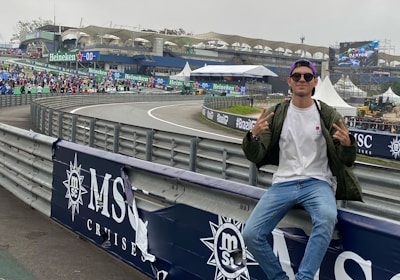 A person in casual attire sits on a barrier beside a racetrack, making peace signs with both hands. In the background, there is a stadium, a large crowd, promotional banners, and a screen displaying an advertisement. The scene has an overcast sky, and the setting appears to be a motorsport event.