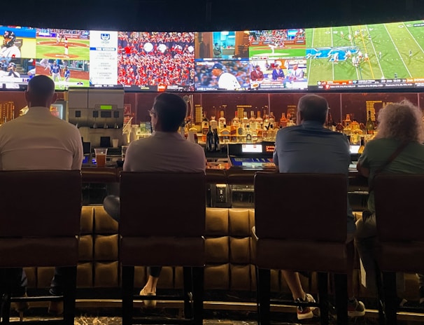 A group of people sit at a bar with their backs to the camera, watching multiple sports games displayed on large screens. The bar counter is lined with various bottles of liquor, and the atmosphere suggests a casual and relaxed social setting.