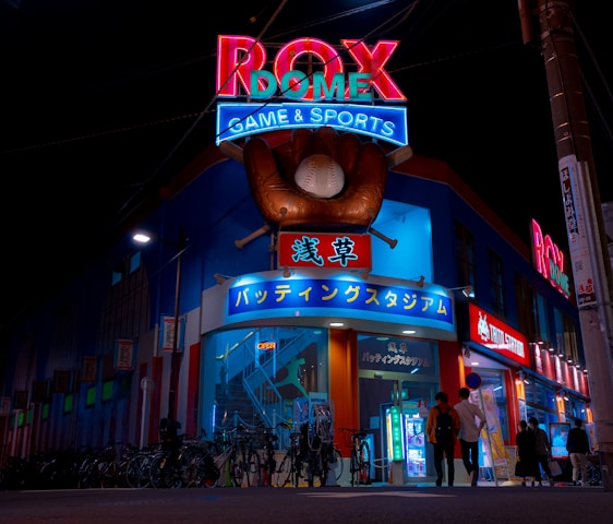 A vibrant, neon-lit facade of a gaming and sports establishment at night, featuring a large glove holding a baseball above the entrance. The building is adorned with colorful signs in both English and Japanese, with various advertisements and an illuminated sign that says 'ROX DOME'. Several people are walking towards the entrance, and bicycles are parked along the side.