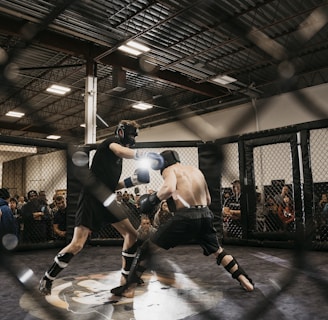 Two fighters engage in a mixed martial arts match within a caged ring. They are wearing protective gear, including gloves and shin guards, and are surrounded by spectators watching intently. Harsh lighting from above casts shadows on the fighters and the mat.